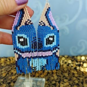 May include: Handmade beaded earrings in a whimsical design. The earrings feature a blue, black, and light blue color scheme, with a fringe at the bottom. The earrings are displayed on a clear stand against a blue background.