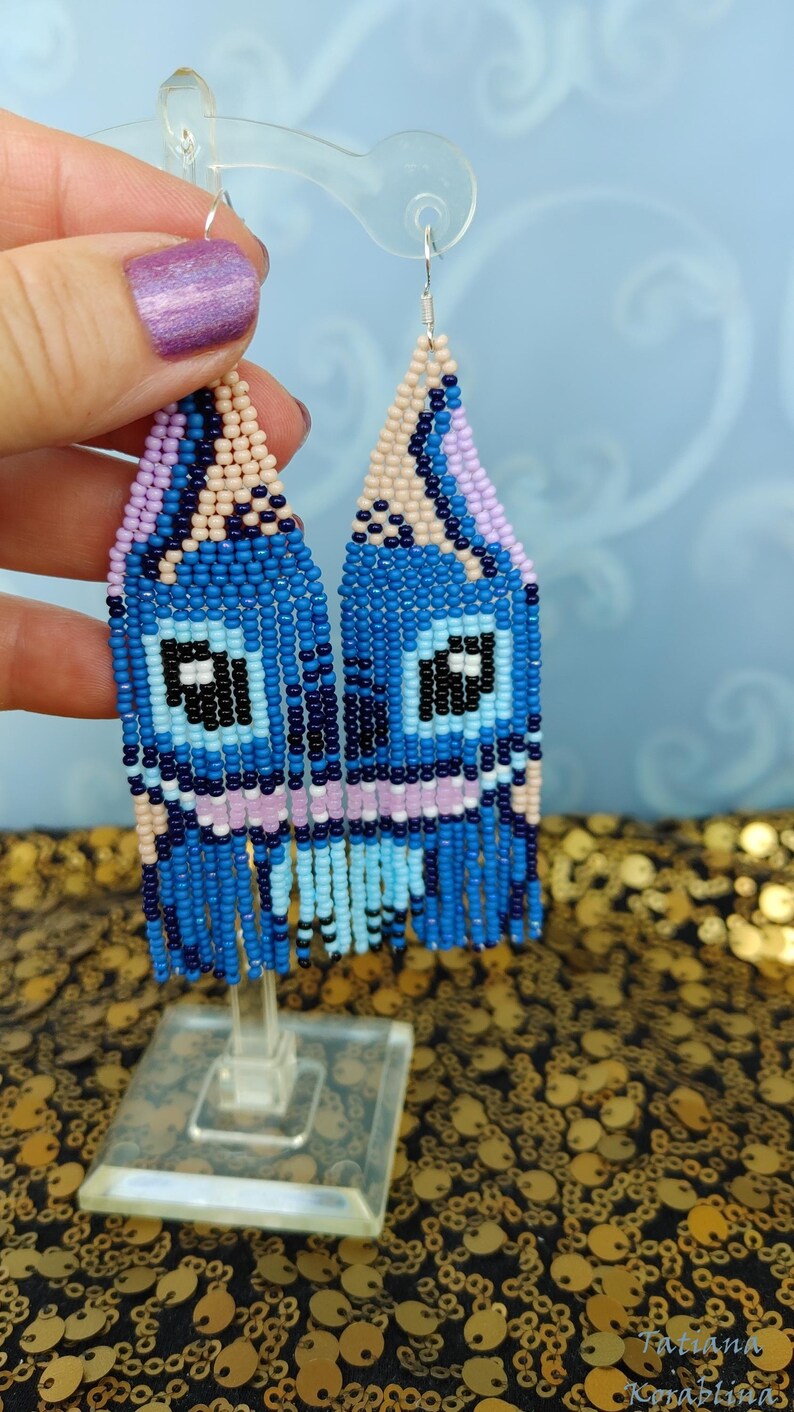 May include: Handmade beaded earrings in a whimsical design. The earrings feature a blue, black, and light blue color scheme, with a fringe at the bottom. The earrings are displayed on a clear stand against a blue background.
