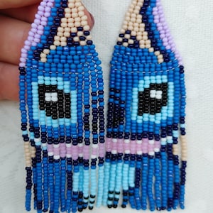 May include: Handmade beaded earrings in a unique design. The earrings feature a blue, black, and white eye-like pattern with dangling blue beads. The top portion is a mix of light pink, beige, and purple beads. The earrings are a striking accessory.