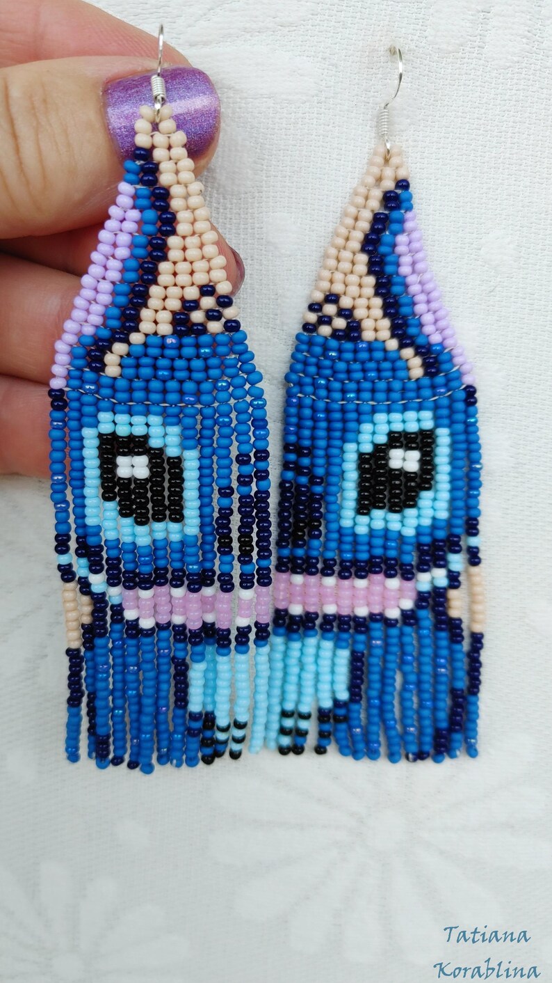 May include: Handmade beaded earrings in a unique design. The earrings feature a blue, black, and white eye-like pattern with dangling blue beads. The top portion is a mix of light pink, beige, and purple beads. The earrings are a striking accessory.