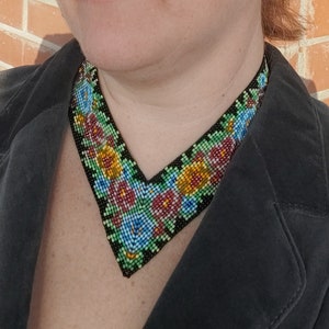 Può includere: Un colletto nero con un design a V decorato con perline floreali colorate. Il colletto è realizzato con piccole perline colorate in tonalità di rosso, blu, verde e giallo.