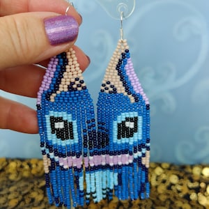 May include: Handmade beaded earrings in shades of blue, light blue, black, pink, and beige. The earrings feature a unique design with a fringe at the bottom. The earrings are displayed on a clear stand. The earrings are a unique accessory.