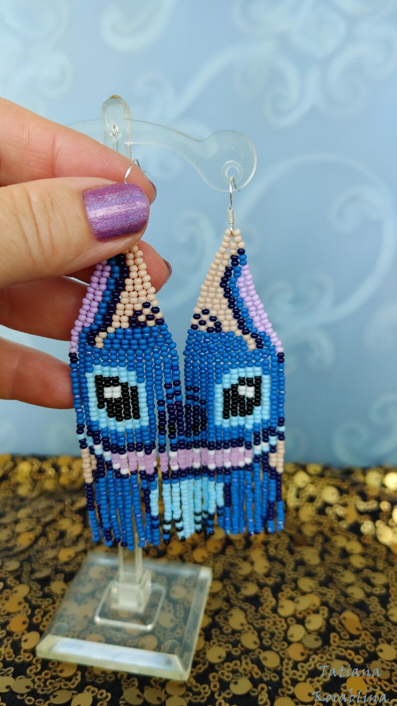 May include: Handmade beaded earrings in shades of blue, light blue, black, pink, and beige. The earrings feature a unique design with a fringe at the bottom. The earrings are displayed on a clear stand. The earrings are a unique accessory.