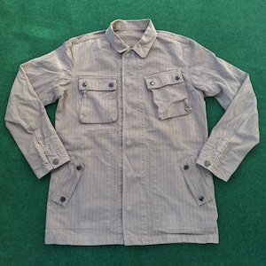 Puede incluir: Chaqueta color beige con estampado de espiga. Tiene cuello, cuatro bolsillos delanteros y cierres de botones. Las mangas tienen puños abotonados. La chaqueta está sobre una superficie verde.