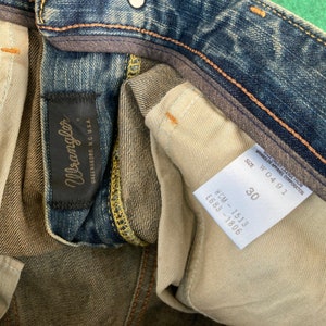 May include: Close-up of a pair of blue denim jeans with a brown leather Wrangler label and a white tag with the text "Wrangler Apparel Corporation" and "Size W0491 30".