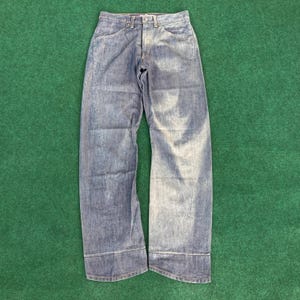 Puede incluir: Un par de vaqueros de mezclilla azul con un diseño clásico de cinco bolsillos. Los vaqueros tienen un corte de pierna recta y un cierre de botón y cremallera. La mezclilla tiene un lavado descolorido, dando a los vaqueros un aspecto desgastado. Los vaqueros están planos sobre una superficie verde.