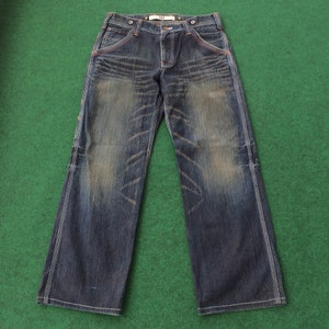 May include: Dark wash denim jeans with a faded effect and white stitching. The jeans have a wide leg cut, orange stitching details, and a button and zipper closure. The jeans are laid flat on a green surface.