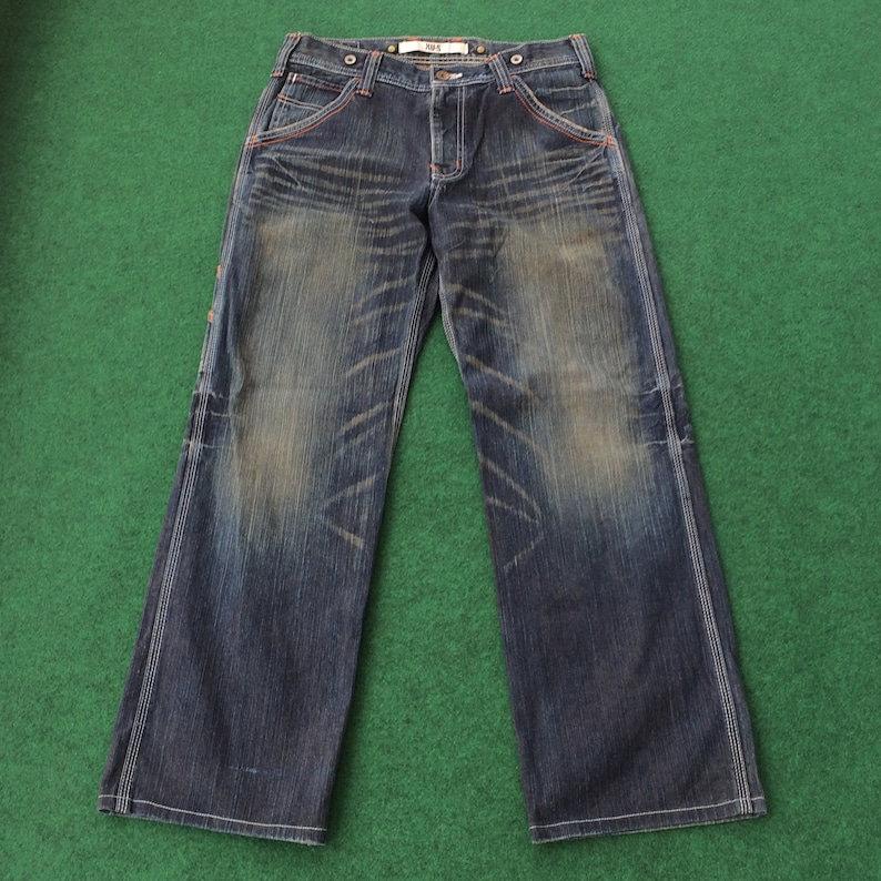 May include: Dark wash denim jeans with a faded effect and white stitching. The jeans have a wide leg cut, orange stitching details, and a button and zipper closure. The jeans are laid flat on a green surface.