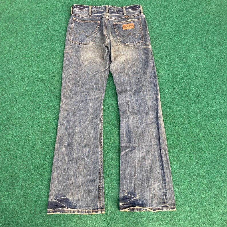 May include: A pair of blue denim jeans with a wide leg opening. The jeans have a faded look and a brown leather patch on the back with the word "Wrangler" printed on it.