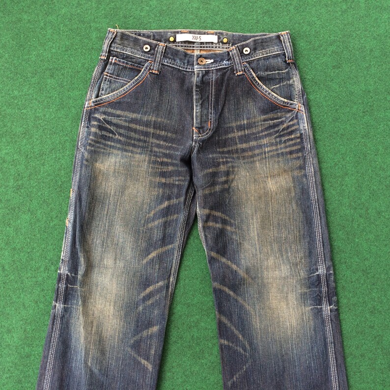 May include: Dark wash denim jeans with a faded, worn appearance. Features orange stitching along the pockets and seams, with the text "XUS" on the waistband. The jeans have a classic five-pocket design and a button and zipper closure.