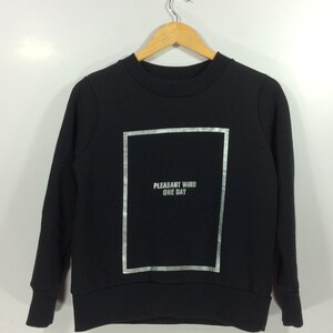 Könnte beinhalten: Schwarzer Pullover mit einem silbernen Grafikdruck eines Rechtecks mit dem Text "PLEASANT WIND ONE DAY" darin.