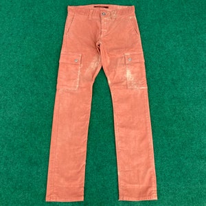 May include: Rust-colored corduroy pants with two cargo pockets on the legs. The pants have a button and zipper closure, and belt loops. The fabric has a slightly faded appearance. The pants are laid flat on a green surface.