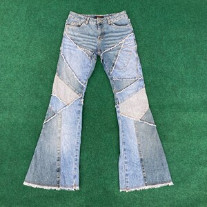 May include: Light blue denim jeans with a patchwork design. The jeans have a high waist and a flared leg. The patchwork is made up of different shades of blue denim, with raw edges. The jeans are laid flat on a green surface.