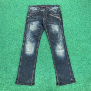 Puede incluir: Vaqueros de mezclilla azul oscuro con un aspecto descolorido y desgastado. Incluyen cremallera, bolsillos delanteros con cremalleras y un diseño clásico de cinco bolsillos. Los vaqueros tienen un corte de pierna recta y están sobre una superficie verde.