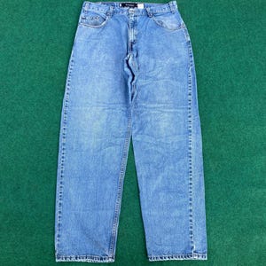 May include: Light blue denim jeans with a classic five-pocket design. The jeans have a straight leg cut and a button and zip closure. The fabric shows subtle fading and wear, giving them a vintage look. The jeans are laid flat on a green surface.