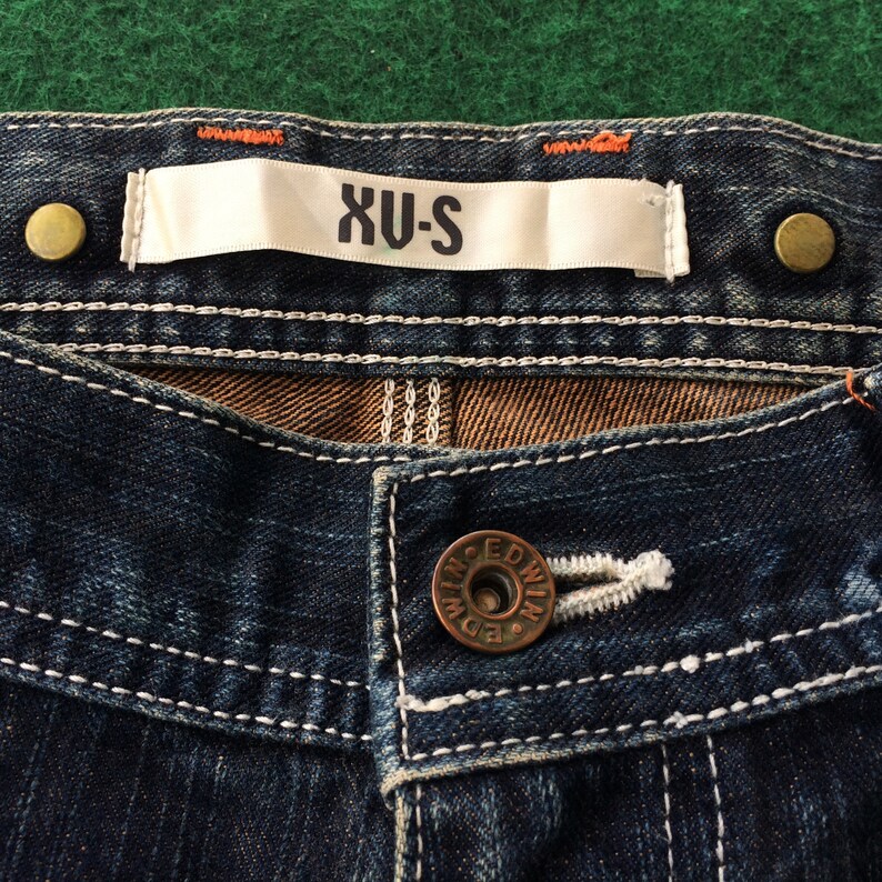May include: Close-up of dark blue denim jeans with white stitching. The jeans feature a button closure with the brand name "EDWIN" and a label that reads "XU-S". Two brass-colored rivets are visible.