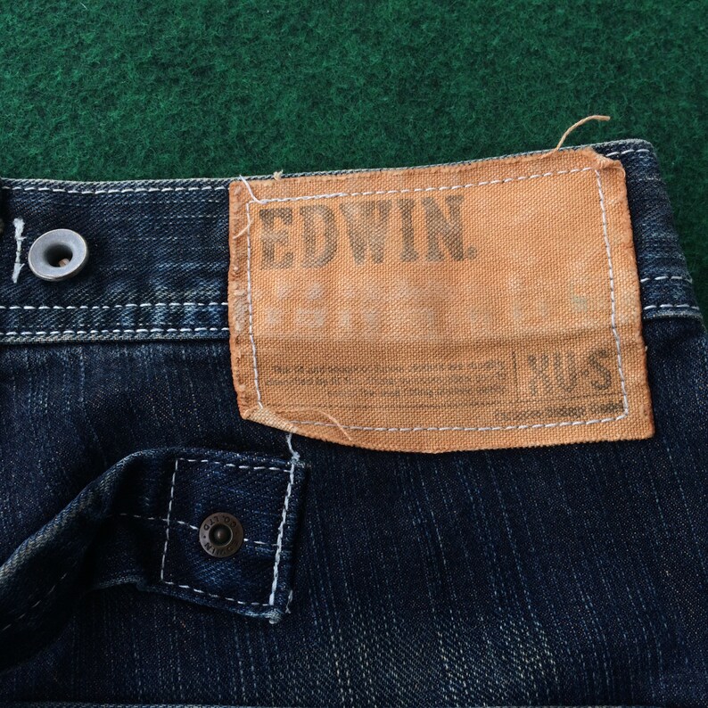 May include: Close-up of dark blue denim fabric with a light brown Edwin label. The label is rectangular with the brand name printed in a dark font. White stitching is visible on the denim and around the label. A metal button is also visible.