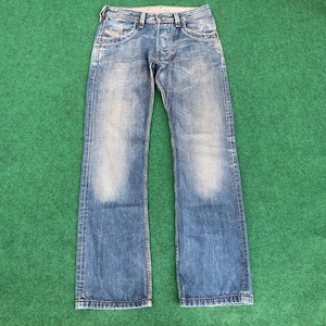 May include: Blue denim jeans with a faded wash and a classic five-pocket design. The jeans have a button fly and a straight leg fit. The fabric shows signs of wear, adding to the vintage aesthetic. The jeans are laid flat on a green surface.