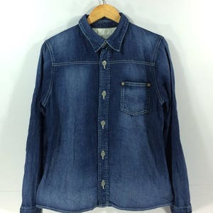 Könnte beinhalten: Ein blaues Jeanshemd mit langen Ärmeln, Knopfleiste und einer Brusttasche.