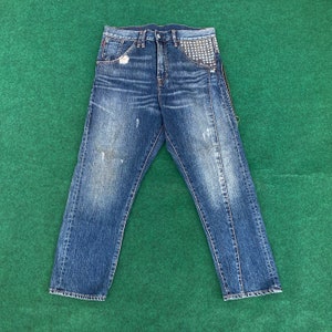 Puede incluir: Vaqueros de mezclilla azul con detalles desgastados y un bolsillo con tachuelas. Los vaqueros tienen una cremallera en la costura lateral.