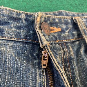 May include: Close-up of the waistband of a pair of blue denim jeans. The jeans have a zipper fly and a metal button with the word "Wrangler" embossed on it.