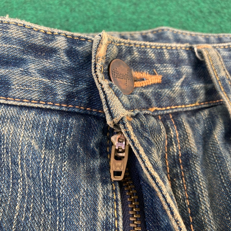May include: Close-up of the waistband of a pair of blue denim jeans. The jeans have a zipper fly and a metal button with the word "Wrangler" embossed on it.
