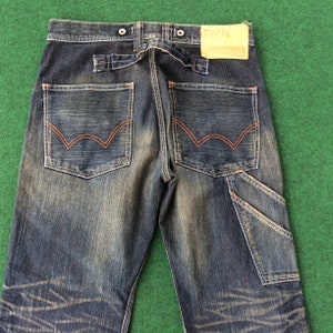 May include: Dark wash denim jeans with orange stitching details. Features include two back pockets with a distinctive design, a utility pocket, and a leather patch. The jeans have a button closure and a belt loop detail.