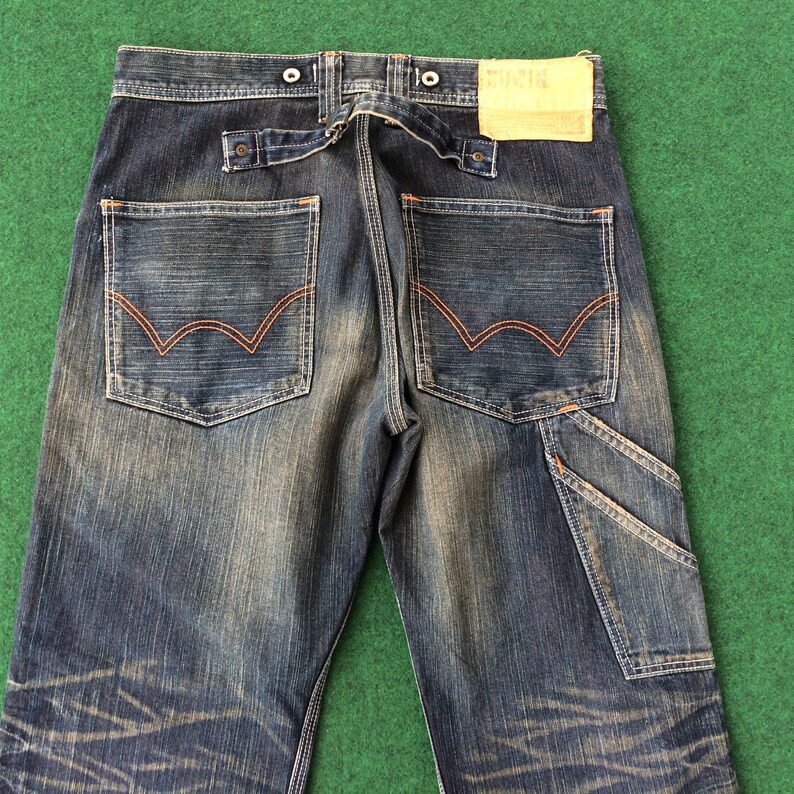 May include: Dark wash denim jeans with orange stitching details. Features include two back pockets with a distinctive design, a utility pocket, and a leather patch. The jeans have a button closure and a belt loop detail.