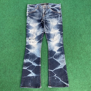 May include: Dark blue denim jeans with a distinctive white pattern and a distressed knee. The jeans feature a classic five-pocket design and a flared leg. The jeans are laid flat on a green surface.