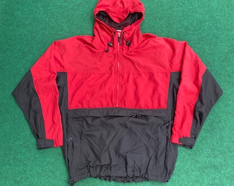 Puede incluir: Una chaqueta tipo anorak roja y negra con capucha. La chaqueta tiene cierre de media cremallera, un bolsillo delantero y un cordón en la cintura. La mitad superior es roja, y la mitad inferior y las mangas son negras.