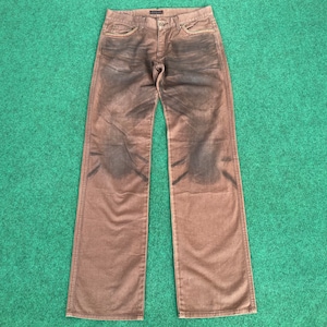 May include: Brown denim jeans with a faded, distressed look. The jeans feature a classic five-pocket design, a button and zip closure, and a straight leg cut. The stitching is visible, and the jeans are laid flat on a green surface.