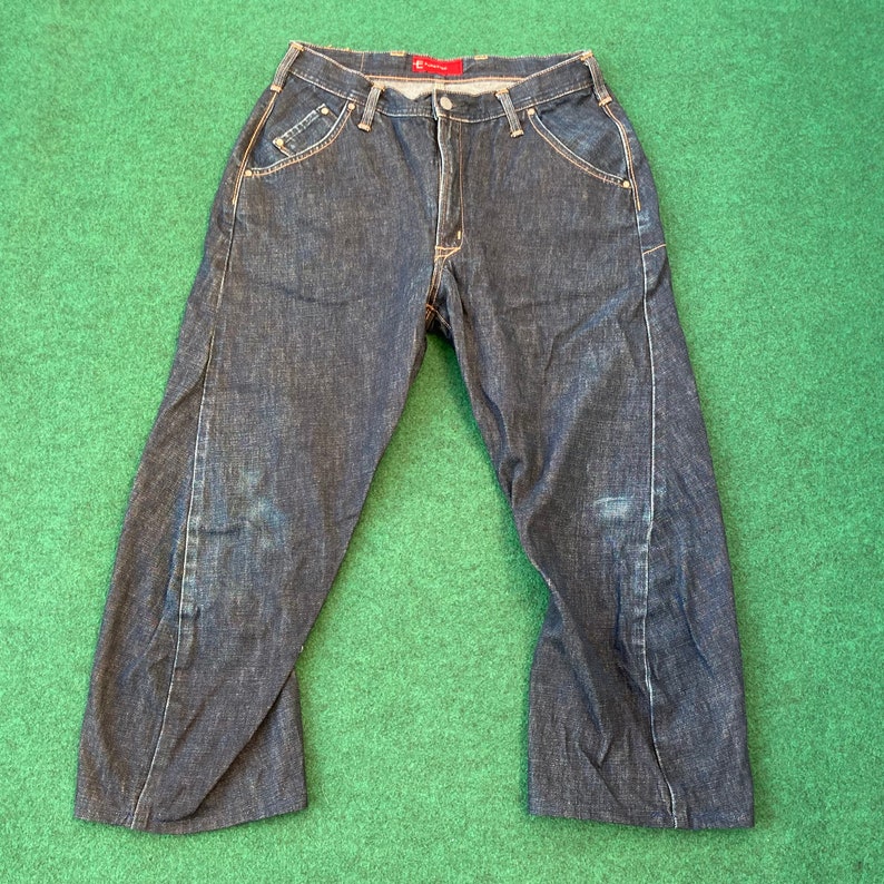 May include: A pair of dark blue denim jeans with a straight leg and a slightly flared hem. The jeans have a red tag on the back waistband.