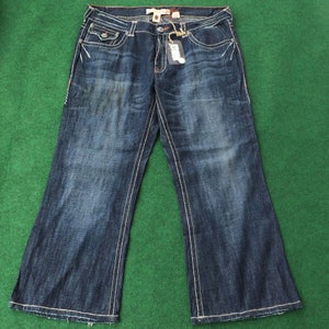 Könnte beinhalten: Dunkelblaue Jeans mit weißen Nähten. Die Jeans haben ein klassisches Fünf-Taschen-Design, einen Knopf- und Reißverschluss und ein Markenetikett. Die Jeans werden auf einer grünen Oberfläche präsentiert.