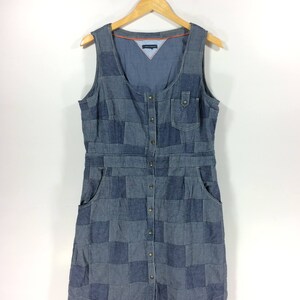 May include: A blue denim dress with a patchwork design. The dress has a sleeveless design, a scoop neck, and a button-up front. The dress has two pockets on the front.