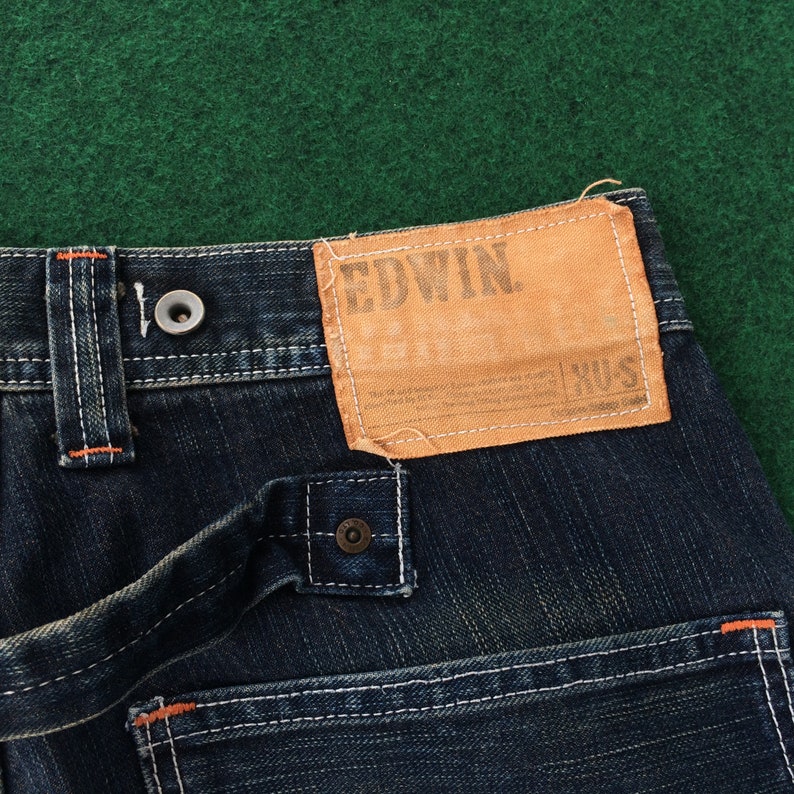 May include: Close-up of dark blue denim jeans with white stitching and orange accents. A tan leather patch with the brand name "EDWIN" is visible. The jeans have a button closure and belt loops.