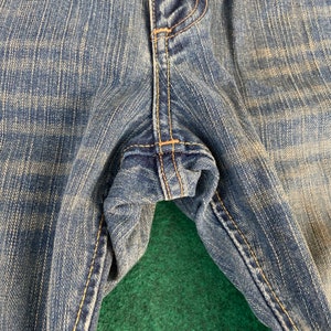 May include: Close-up of the seam of a pair of blue denim jeans. The fabric is a light wash with a faded appearance.
