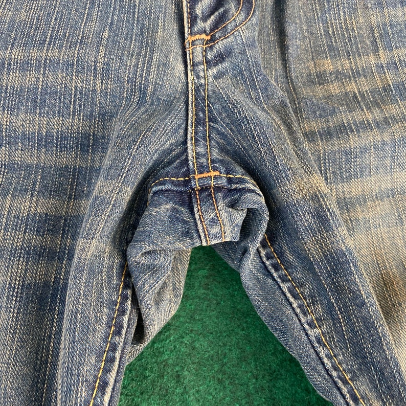 May include: Close-up of the seam of a pair of blue denim jeans. The fabric is a light wash with a faded appearance.