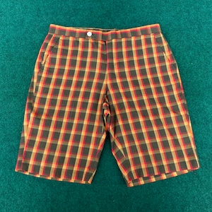May include: A pair of plaid shorts with a button closure and pockets. The shorts have a brown, red, and gold plaid pattern. The garment is displayed on a green surface. The shorts are a classic style.