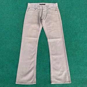 Puede incluir: Pantalones vaqueros de color marrón claro con cierre de botones y diseño de cinco bolsillos. Los vaqueros tienen un corte recto clásico y están hechos de un material de mezclilla duradero. Los vaqueros están colocados sobre una superficie verde.