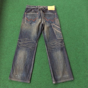May include: Dark wash denim jeans with a faded, distressed look. Features include red stitching on the back pockets, a carpenter pocket, and a leather patch. The jeans have a wide leg and a button closure.