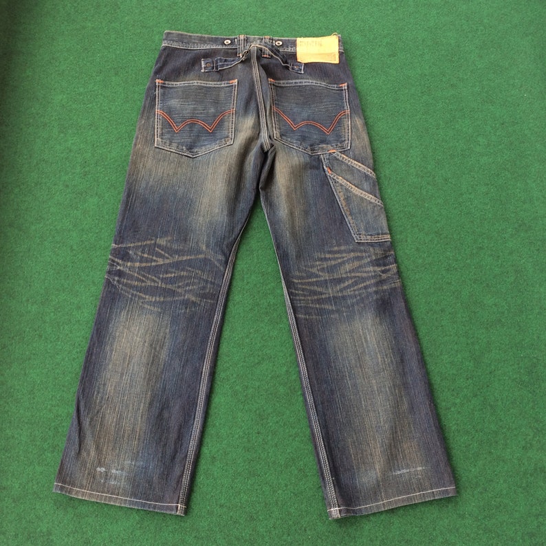 May include: Dark wash denim jeans with a faded, distressed look. Features include red stitching on the back pockets, a carpenter pocket, and a leather patch. The jeans have a wide leg and a button closure.