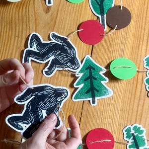 May include: A handmade woodland-themed garland featuring black animal silhouettes, red mushrooms, green trees, and red and green circles strung on twine. The garland is designed for festive decorations.