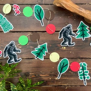 May include: A whimsical garland featuring cut-out shapes of Bigfoot, trees, mushrooms, and circles in green, red, brown, and yellow. The string is made of natural twine, perfect for a woodland-themed party or nursery decoration.
