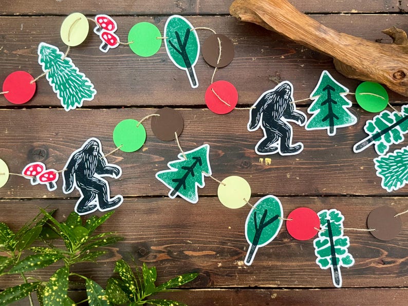 May include: A whimsical garland featuring cut-out shapes of Bigfoot, trees, mushrooms, and circles in green, red, brown, and yellow. The string is made of natural twine, perfect for a woodland-themed party or nursery decoration.