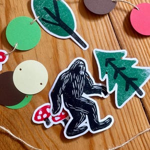 May include: A whimsical garland featuring forest-themed cutouts. Includes a black and white Bigfoot, red and white mushrooms, green leaves, and a green tree. The garland is strung with twine and has various colored circular cutouts.