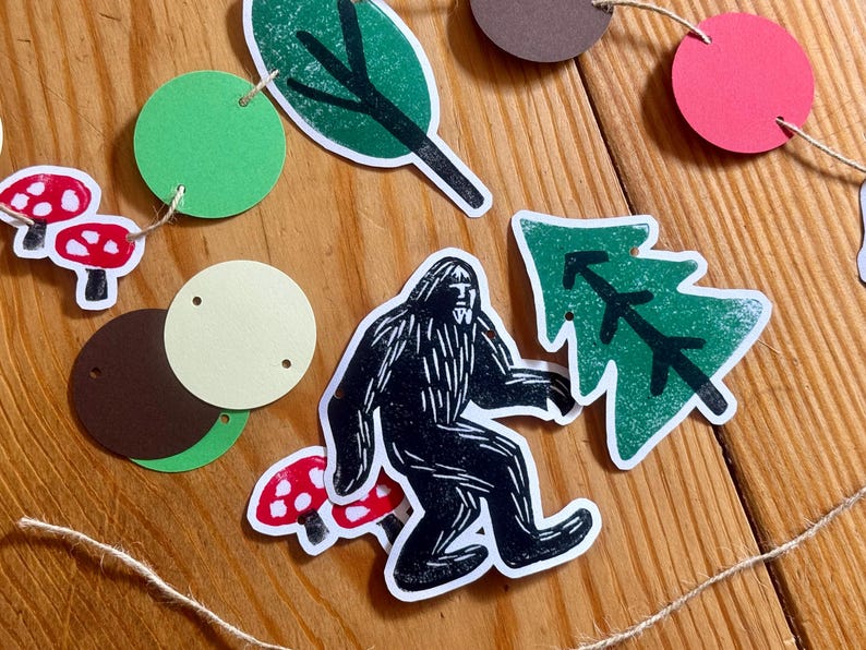 May include: A whimsical garland featuring forest-themed cutouts. Includes a black and white Bigfoot, red and white mushrooms, green leaves, and a green tree. The garland is strung with twine and has various colored circular cutouts.