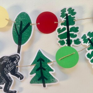 May include: A whimsical garland featuring cut-out shapes of trees, circles, and a Bigfoot silhouette. The shapes are in shades of green, red, yellow, and brown, strung together with twine. This decorative banner is perfect for a woodland-themed party or nursery.