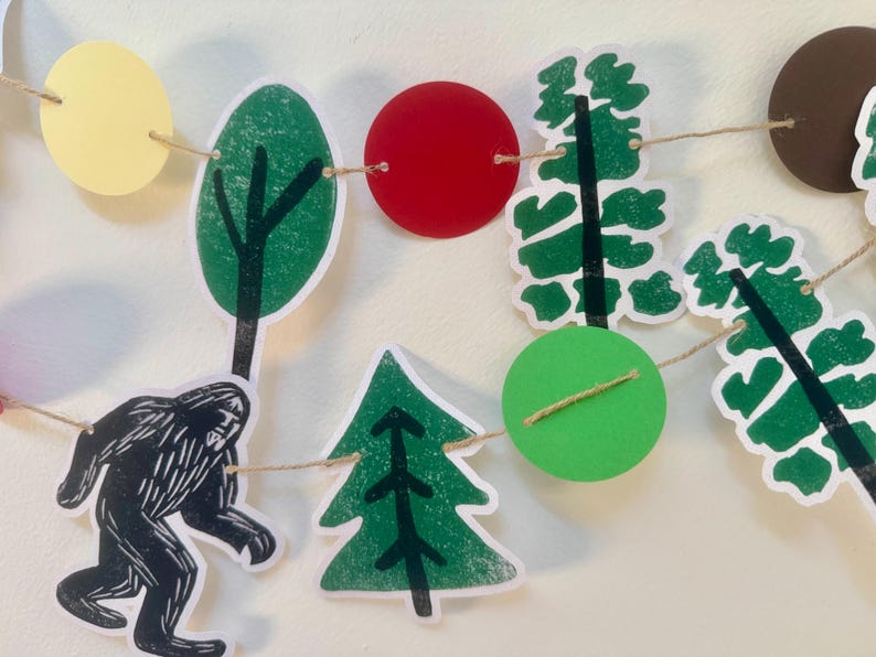 May include: A whimsical garland featuring cut-out shapes of trees, circles, and a Bigfoot silhouette. The shapes are in shades of green, red, yellow, and brown, strung together with twine. This decorative banner is perfect for a woodland-themed party or nursery.