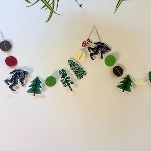 May include: A whimsical garland featuring woodland-themed cutouts. The garland includes black silhouettes of a creature, green trees, red and brown circles, and green leaves. The elements are strung together with twine, creating a decorative accent.