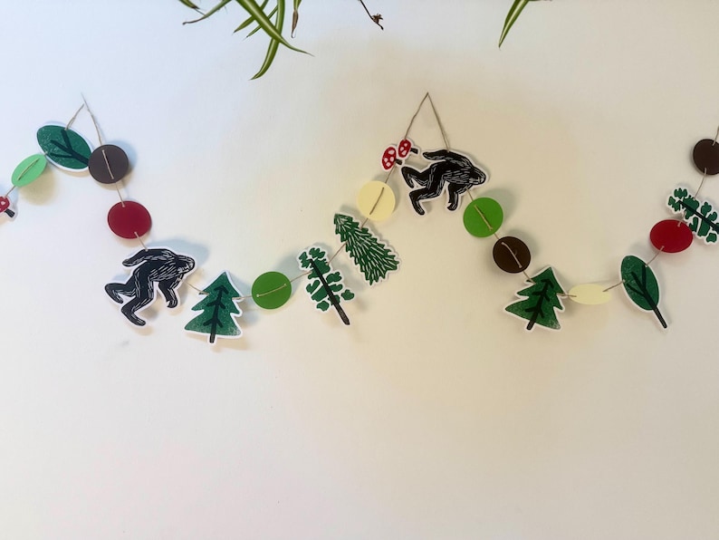May include: A whimsical garland featuring woodland-themed cutouts. The garland includes black silhouettes of a creature, green trees, red and brown circles, and green leaves. The elements are strung together with twine, creating a decorative accent.